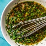 Close up of a whisk in a bowl of chimichurri sauce set on teal napkins with a text overlay on the photo.