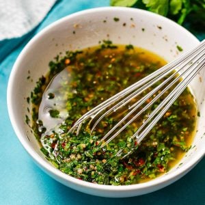 Close up of a whisk resting in a bowl of homemade chimichurri sauce.