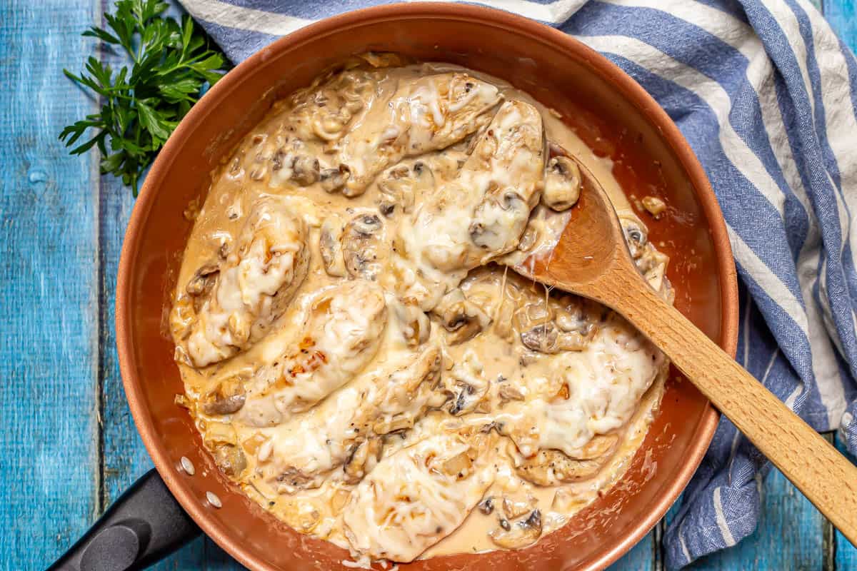 A wooden spoon resting in a large copper skillet with a creamy mixture coating chicken strips and some mushrooms and onions.