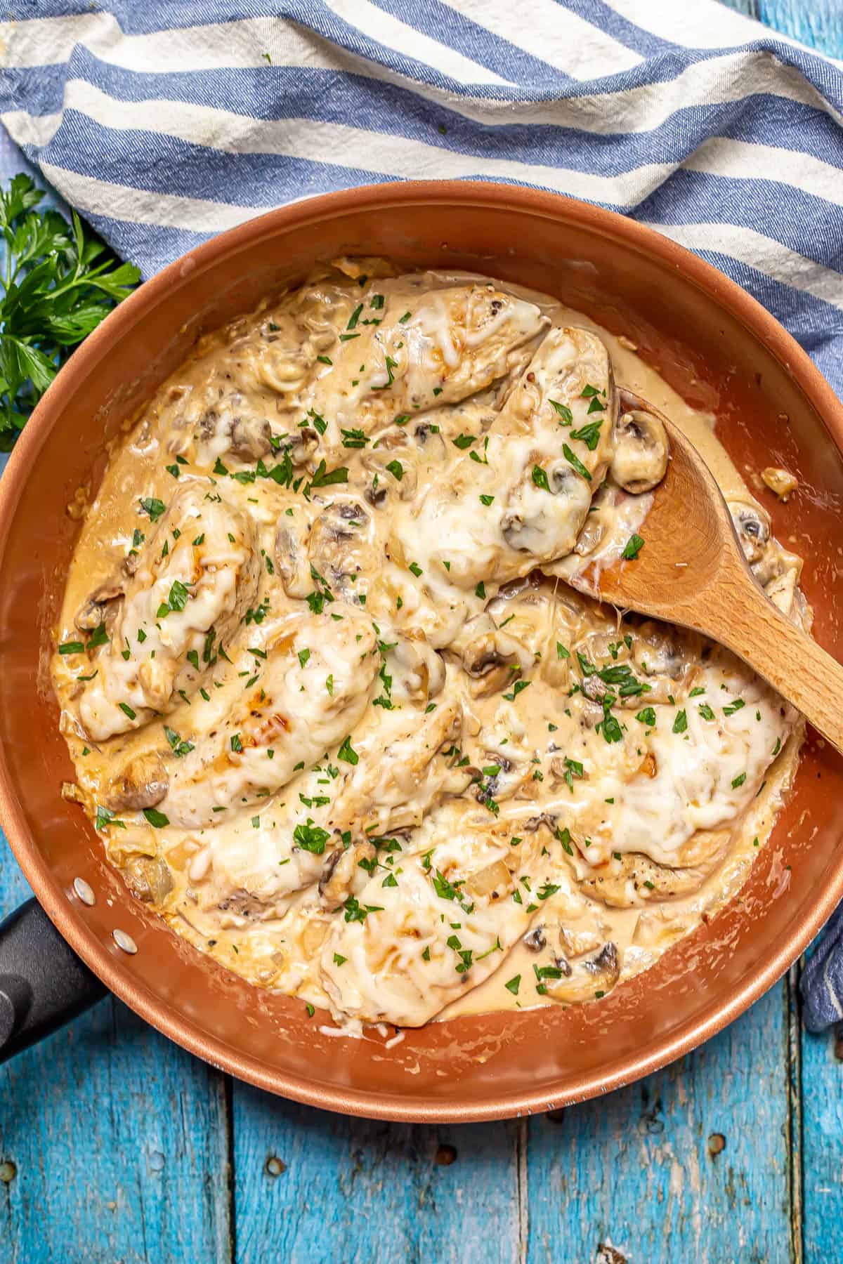 Cream cheese chicken cooked in a copper skillet with a wooden spoon resting in the pan and chopped fresh parsley sprinkled on top.