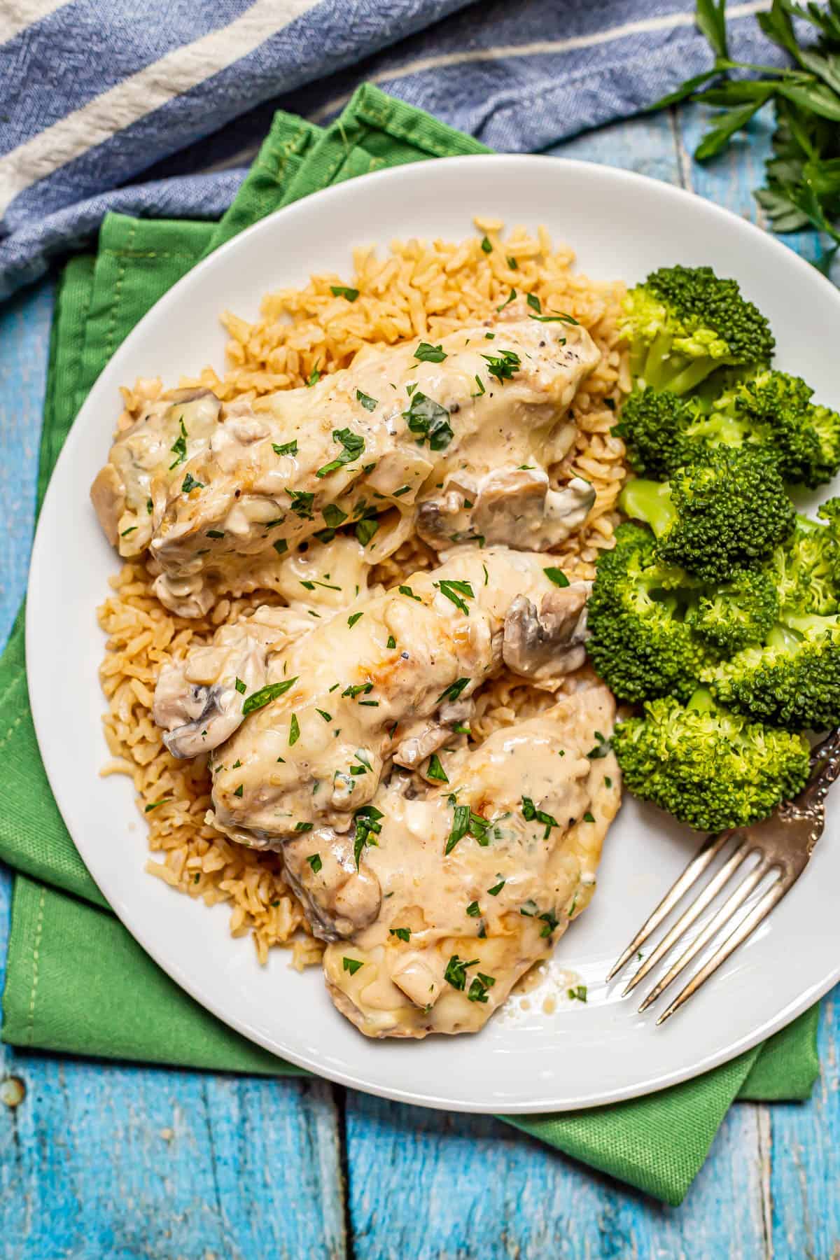 A white dinner plate with cream cheese chicken served over brown rice with steamed broccoli to the side.