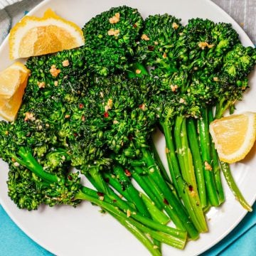Broccolini spears that are sautéed with garlic and red pepper flakes and served with lemon wedges on a white round plate.