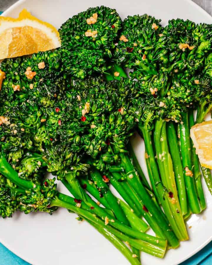 Broccolini spears that are sautéed with garlic and red pepper flakes and served with lemon wedges on a white round plate.