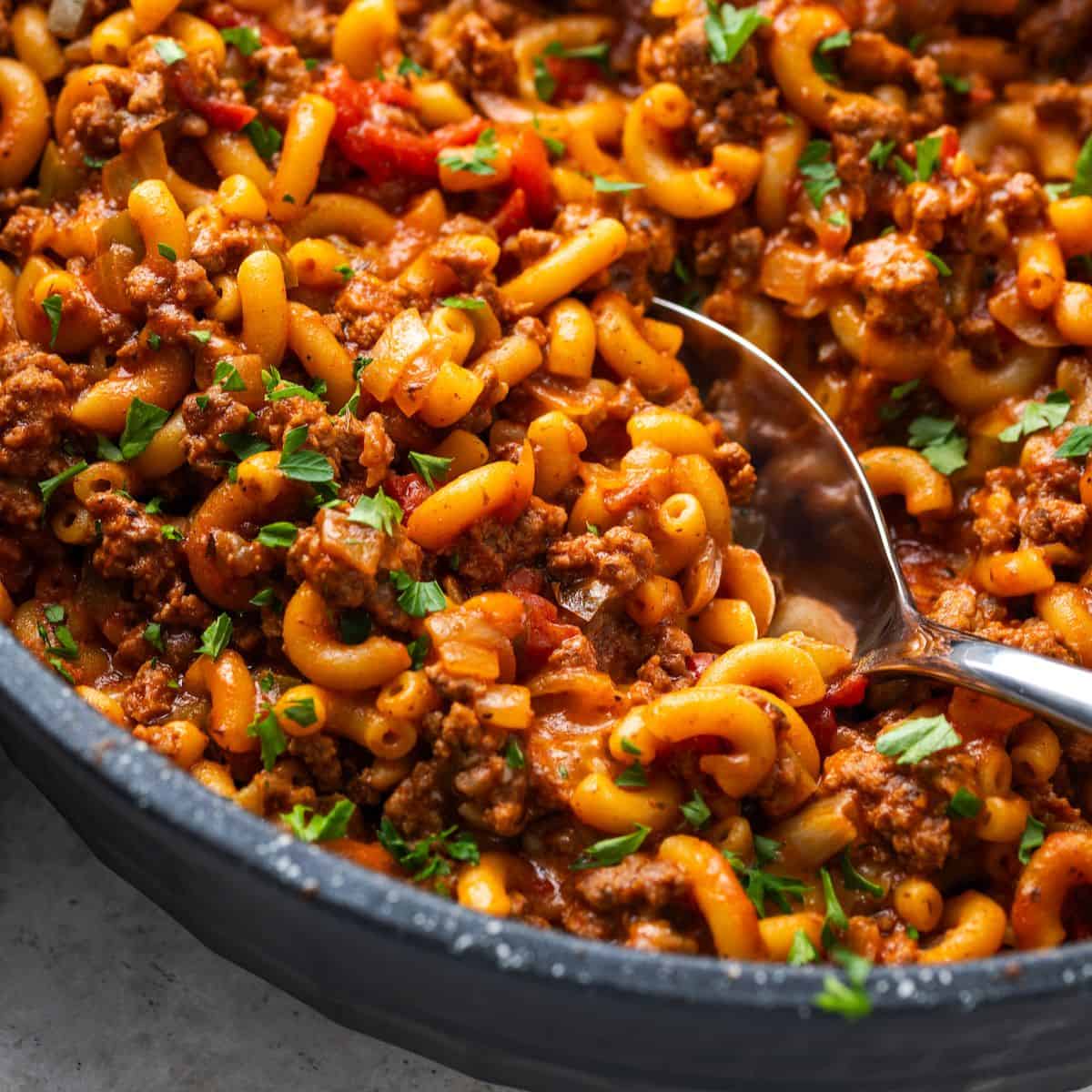 Close up of a spoon scooping up some goulash from a large skillet.