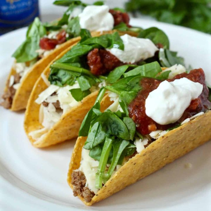 Three hard shelled ground turkey tacos served on a white plate with cheese, spinach, salsa and sour cream as toppings.