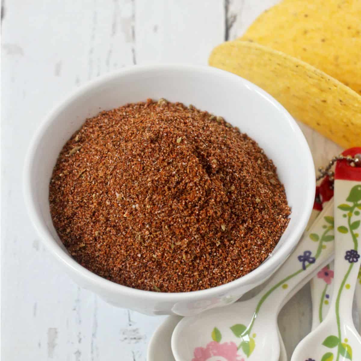 A small white bowl with a homemade taco seasoning mix beside measuring spoons and a hard taco shell.