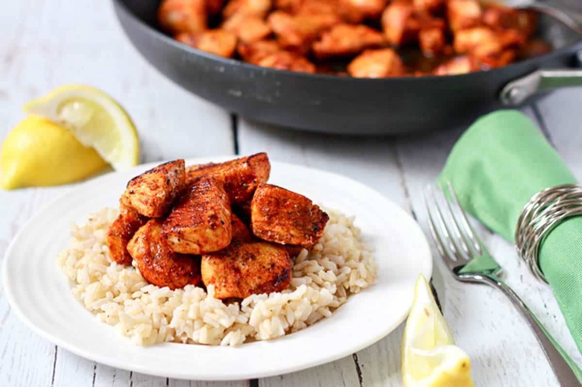 A small white plate with rice topped with sauteed paprika chicken with lemon wedges nearby and a fork to the side.