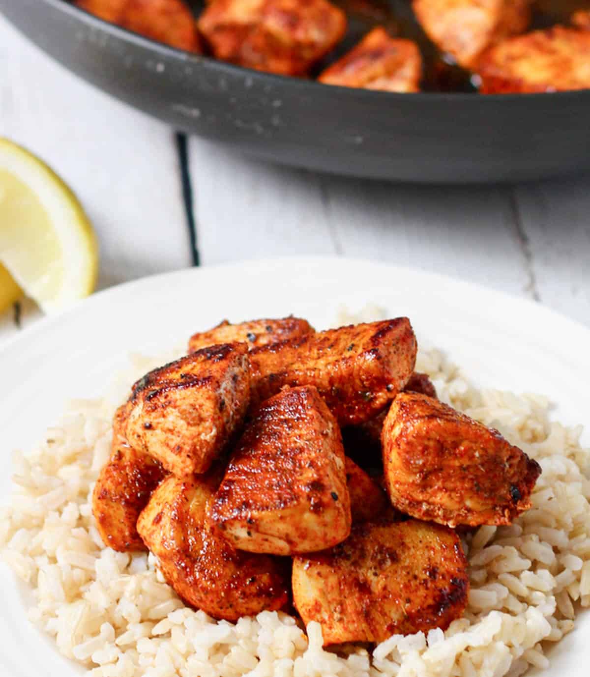 Close up of a small white round plate with fluffy steamed brown rice topped with seared paprika chicken cubes with the skillet in the background.