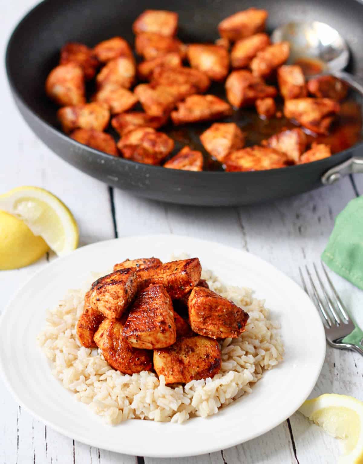 A small white round plate with fluffy steamed brown rice topped with seared paprika chicken cubes with the skillet in the background.