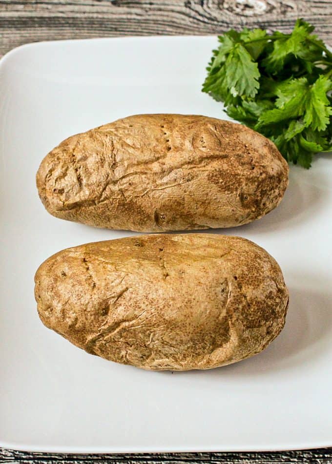 Two potatoes cooked in the microwave on a rectangular white platter with fresh herbs to the side.