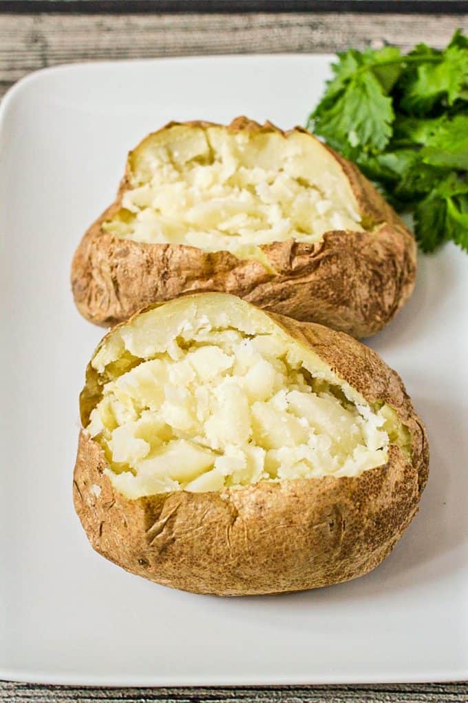 Two potatoes cooked in the microwave cut open and fluffed and served on a rectangular white platter with fresh herbs to the side.