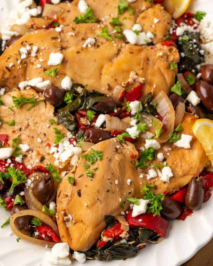 Close up of a white platter with slow cooker Greek chicken with olives, peppers and onions topped with feta cheese and fresh herbs.