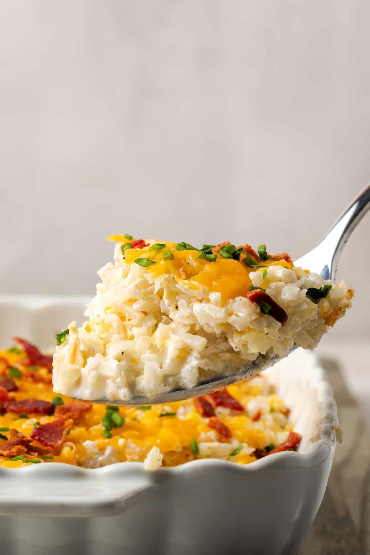 A spoon scooping up some baked cheesy cauliflower rice from a white casserole dish.