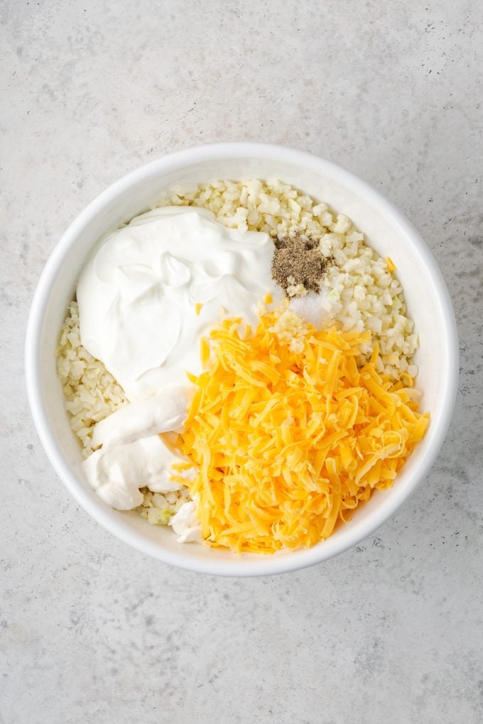 A white mixing bowl with cauliflower rice topped with cheese and Greek yogurt and seasonings before being stirred together.