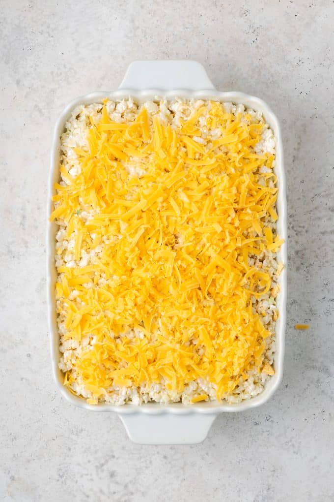 Shredded cheese on top of a creamy cauliflower rice mixture in a white casserole dish before being baked.