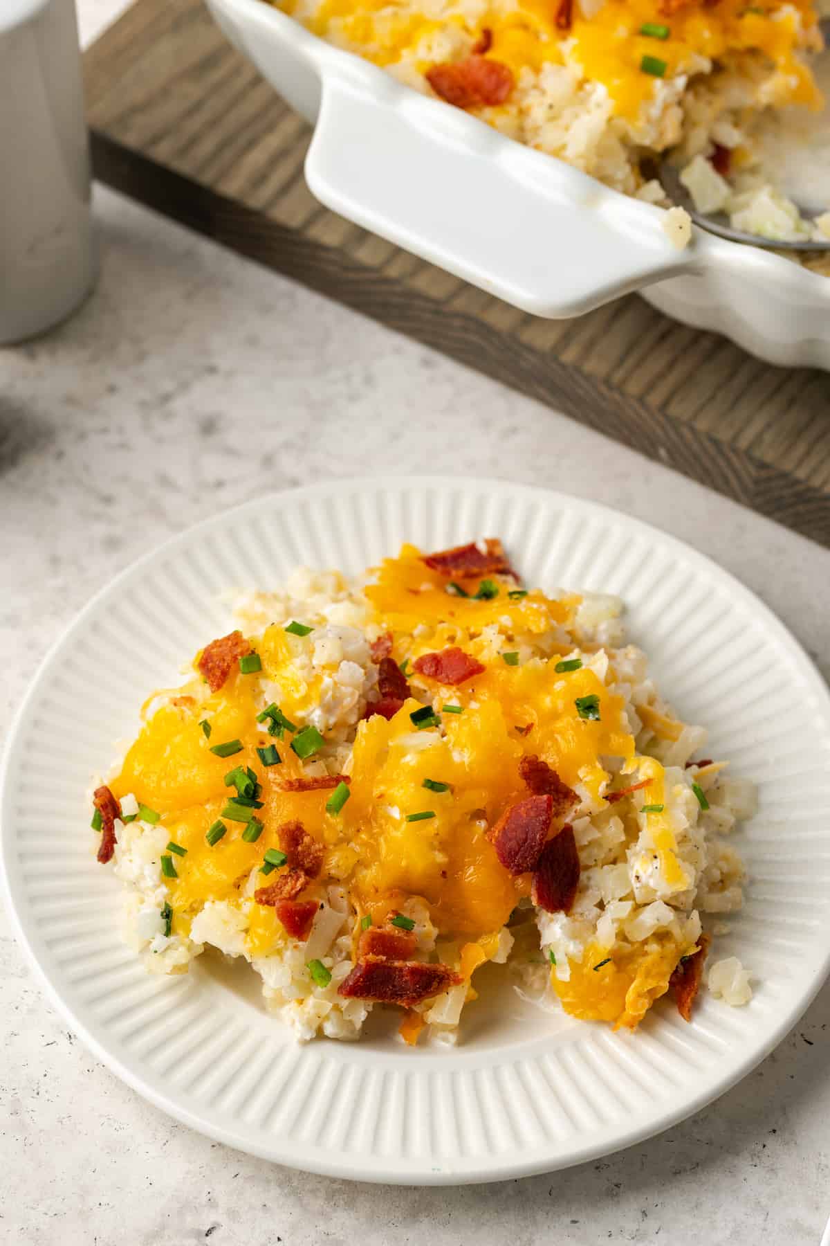 A serving of cheesy cauliflower rice served on a white plate with bacon and chives sprinkled on top.