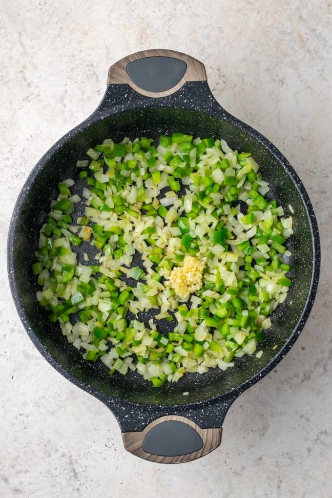 A skillet with sautéed onion and green pepper and garlic being added.