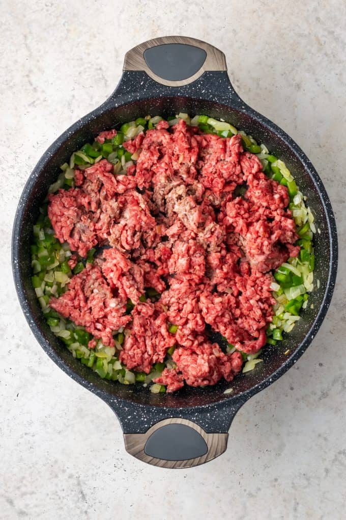 A skillet with sautéed onion and green pepper and ground beef being added.