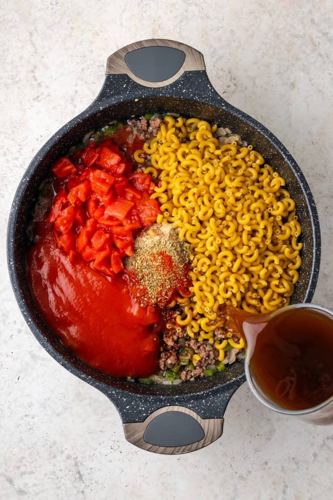 A skillet mixture of ground beef with elbow pasta, tomatoes, tomato sauce, seasonings and broth being added.