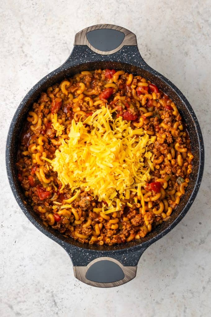 A cooked skillet mixture of ground beef and elbow pasta in a tomato sauce with shredded cheese added on top before being stirred in.