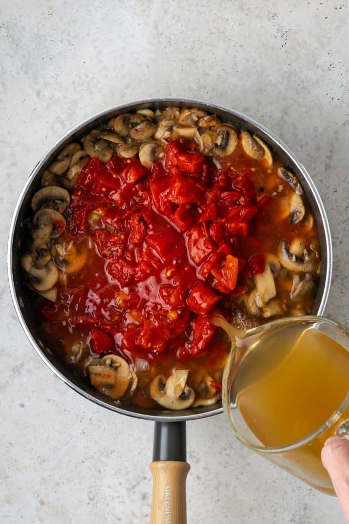 Broth being poured over diced tomatoes on top of mushrooms in a skillet.