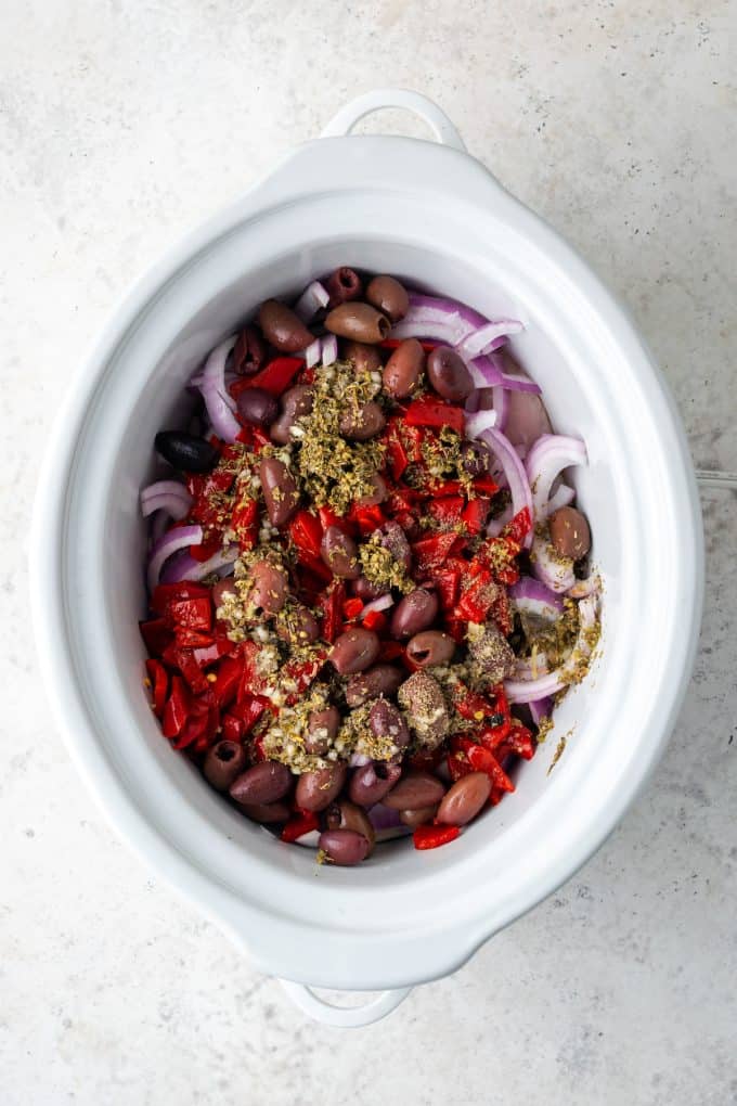 A slow cooker with layers of chicken, red onion, roasted red peppers and olives with an herby sauce poured over everything.