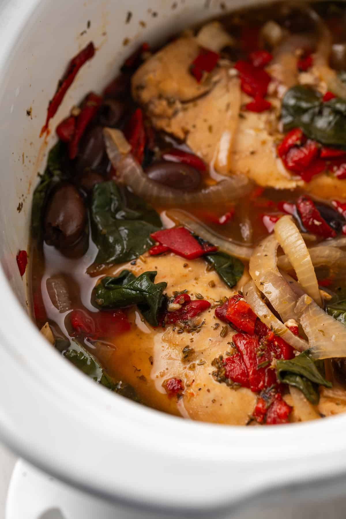 Close up of slow cooker Greek chicken with onions, tomatoes, olives and spinach in the crock pot.