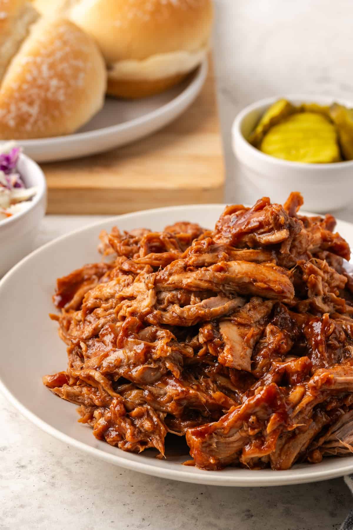 A plate of saucy pulled pork with pickles, coleslaw and burger buns in the background.