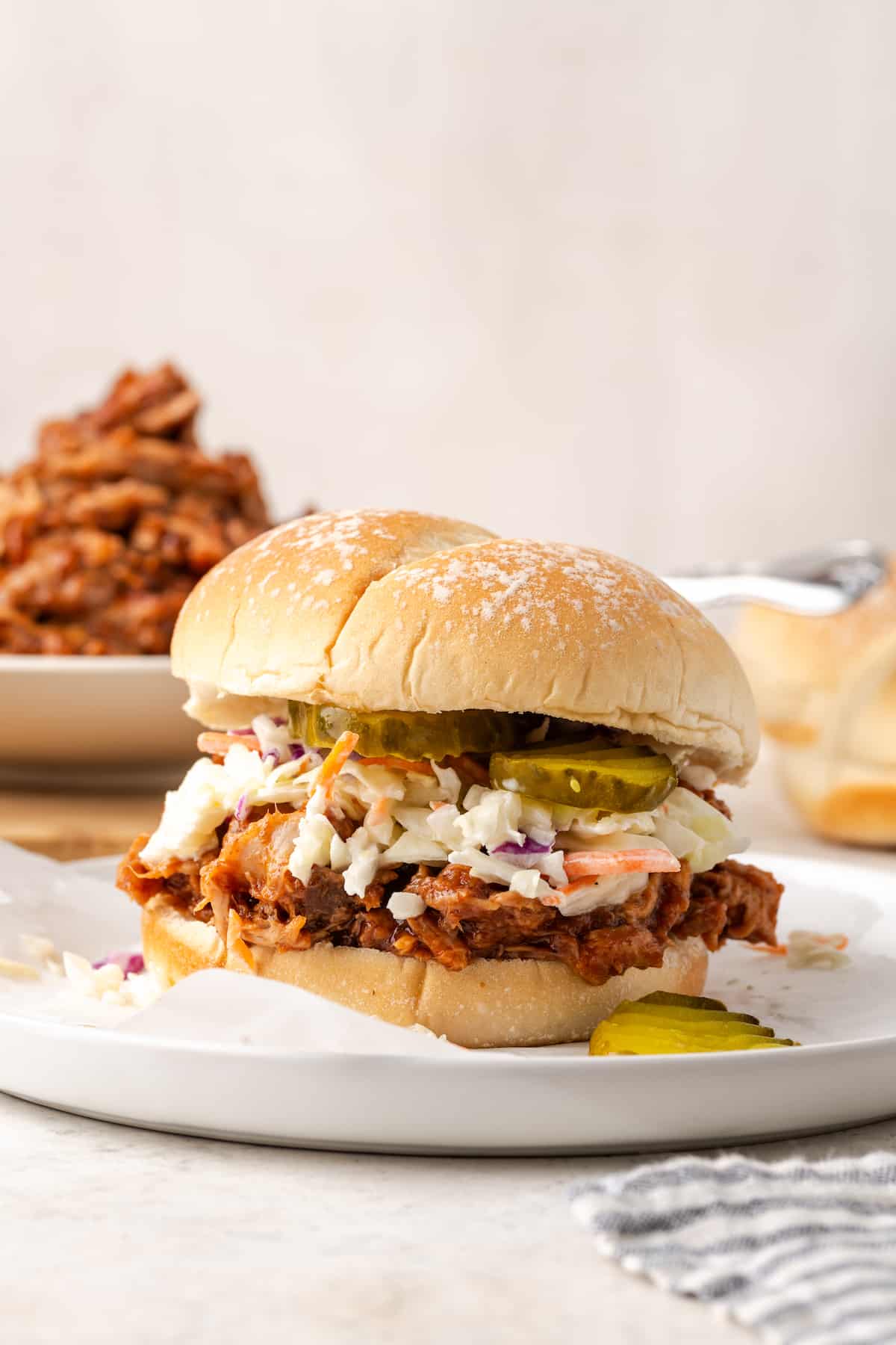 A white plate with a pulled pork sandwich topped with coleslaw and pickles.