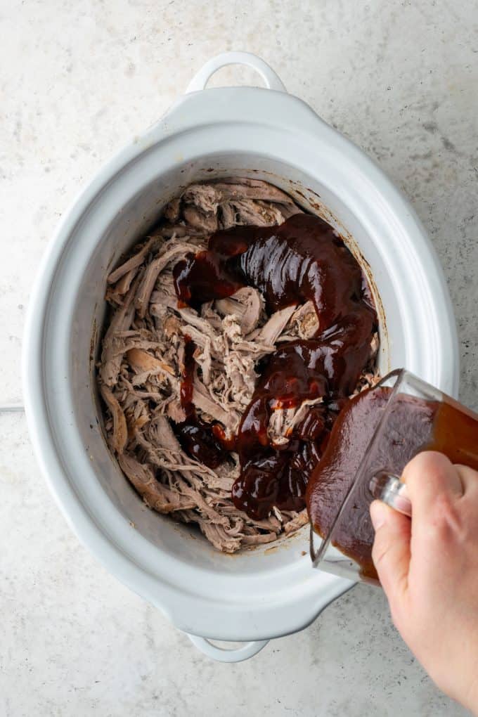 BBQ sauce being poured over cooked, shredded pulled pork in a slow cooker.