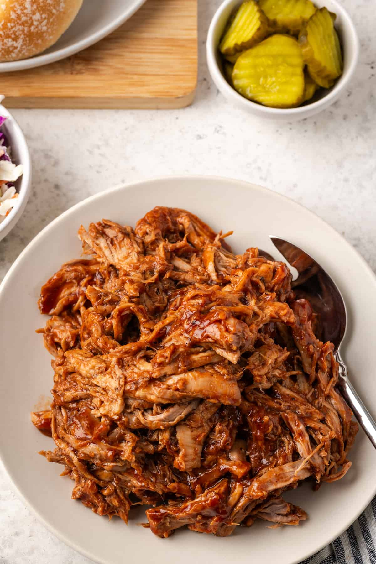 A silver serving fork resting on a plate of pulled pork with a bowl of pickles to the side.