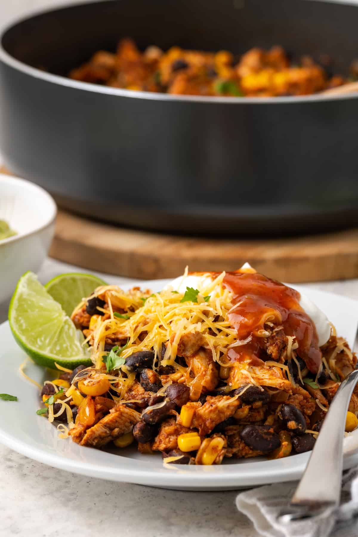 A serving of a southwest chicken and black bean mixture on a white plat with cheese, sour cream and salsa on top and the skillet in the background.