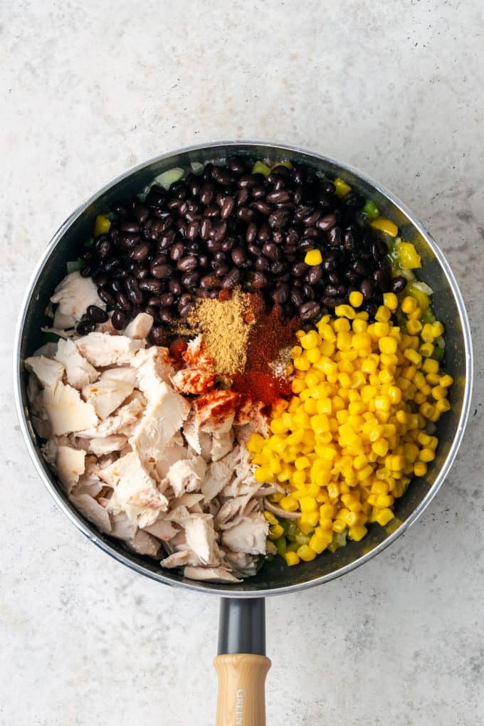 A skillet with sections of chicken, black beans, corn and seasonings being added before being stirred together.