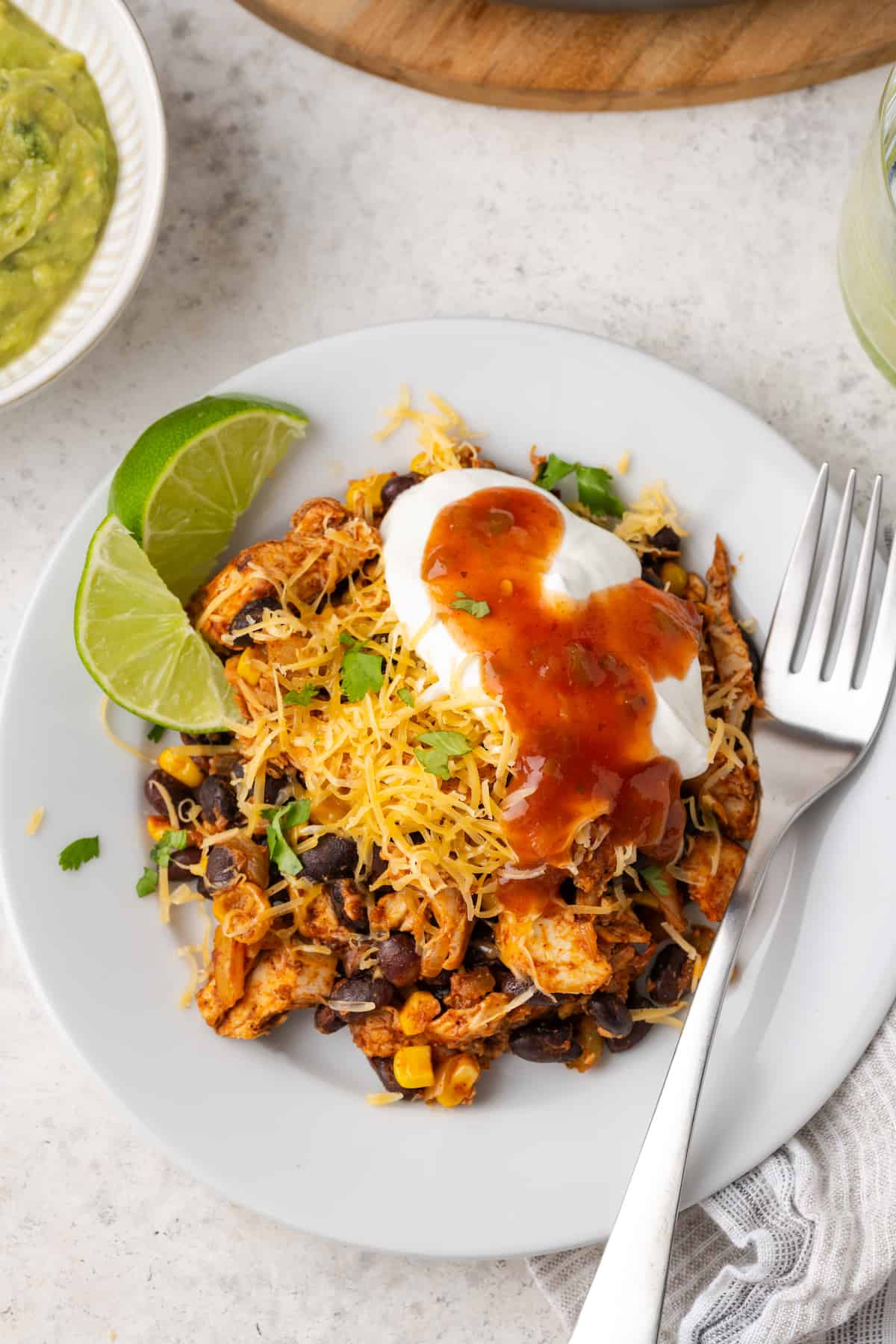 A serving of a southwest chicken and black bean mixture on a white plat with cheese, sour cream and salsa on top and lime wedges and a fork to the side.