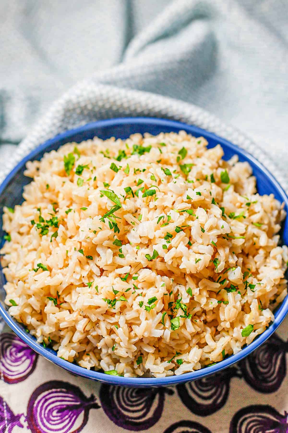 Fluffy steamed brown rice in a blue bowl with chopped fresh parsley on top and colored throw towels to the side.