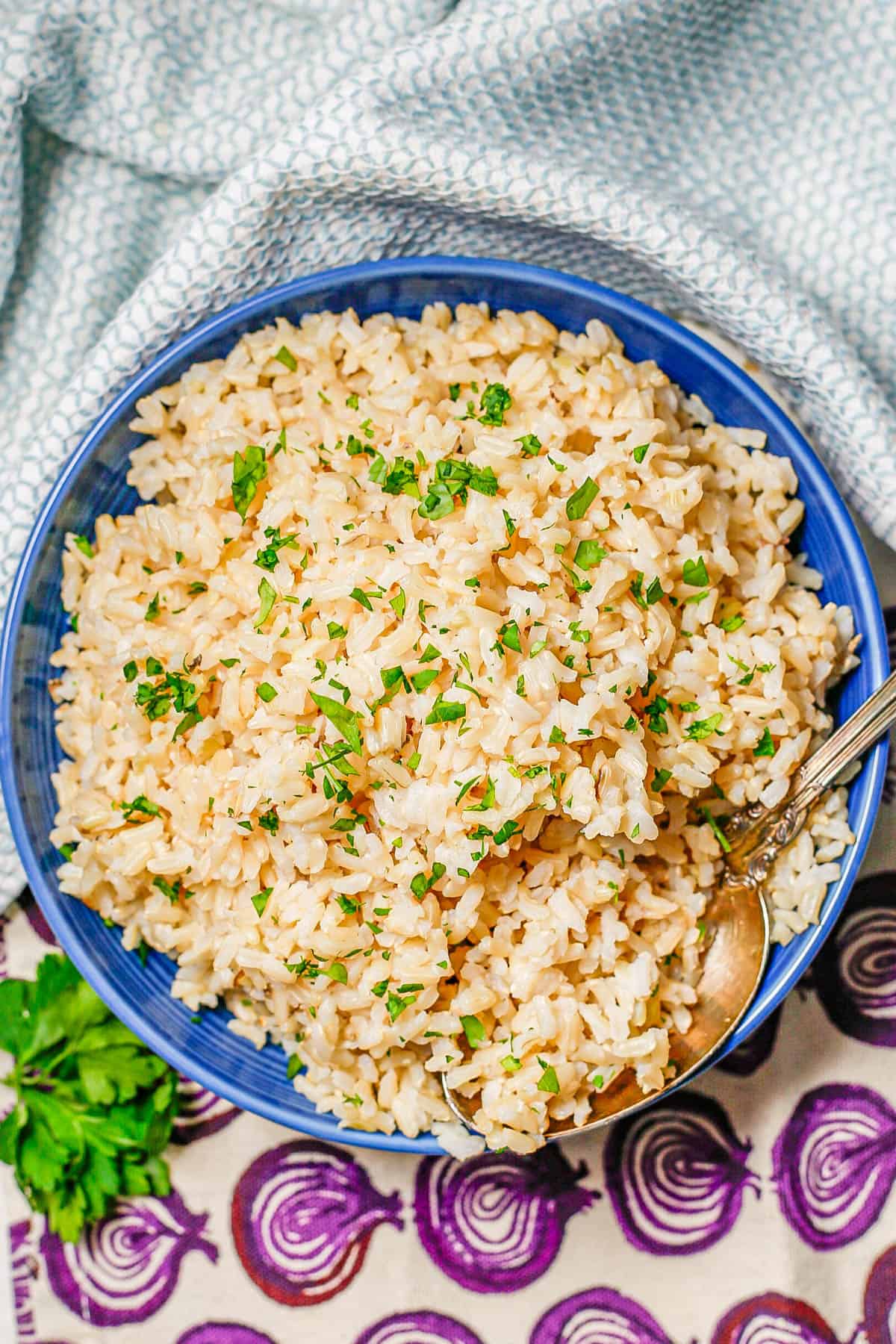 A spoon resting in a blue bowl of fluffy steamed brown rice with chopped fresh parsley on top.