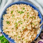 A spoon resting in a blue bowl of fluffy steamed brown rice with chopped fresh parsley on top with a text overlay on the photo.