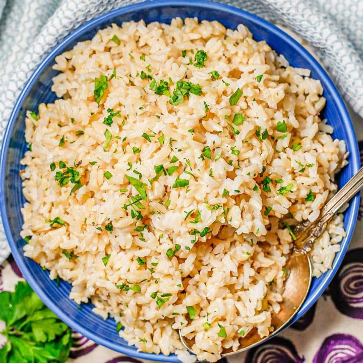 Close up of a spoon resting in a blue bowl of fluffy steamed brown rice with chopped fresh parsley on top.