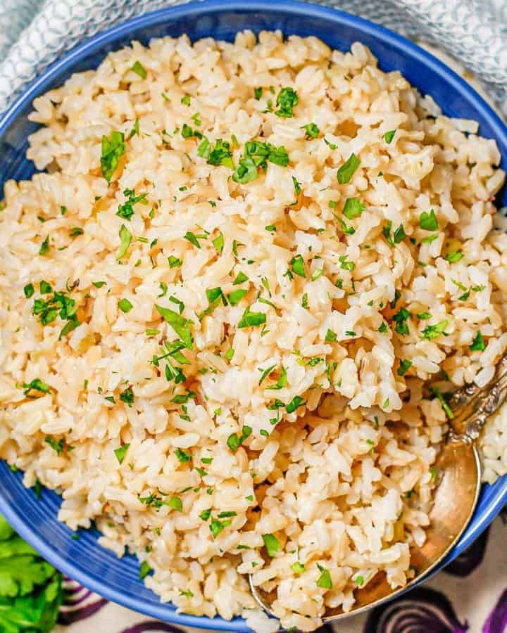 Close up of a spoon resting in a blue bowl of fluffy steamed brown rice with chopped fresh parsley on top.