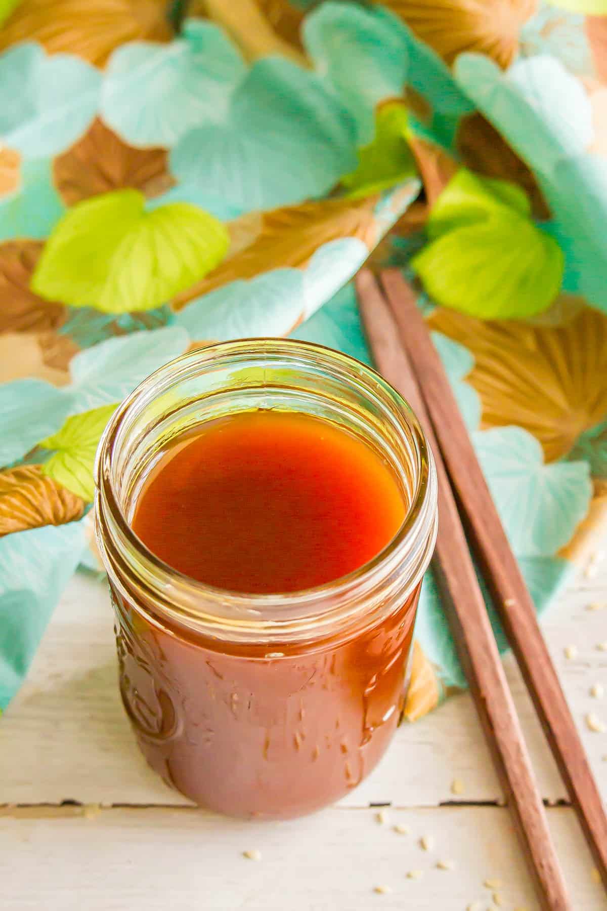 An angled view of an open glass jar with homemade sweet and sour sauce beside a throw napkin and some wooden chopsticks.