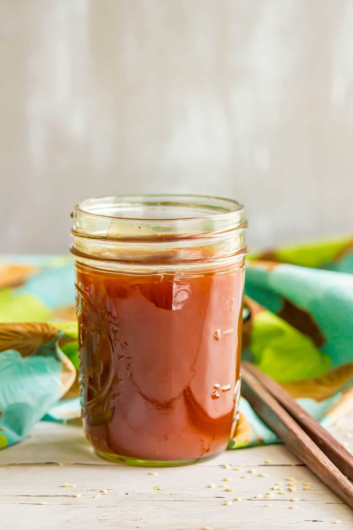 A glass jar filled with a sweet and sour sauce for cooking alongside some sprinkled sesame seeds and a pair of chopsticks.