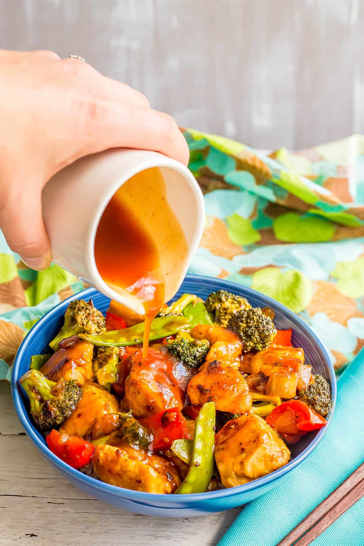 A hand pouring sweet and sour sauce over a bowl full of charred chicken pieces and a mix of vegetables.