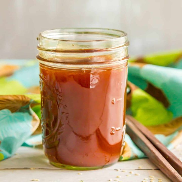 Close up of a glass jar filled with a sweet and sour sauce for cooking alongside some sprinkled sesame seeds and a pair of chopsticks.