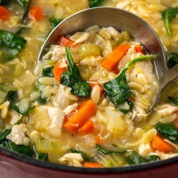 Close up of a silver ladle scooping up lemony chicken orzo soup with spinach from a large pot.