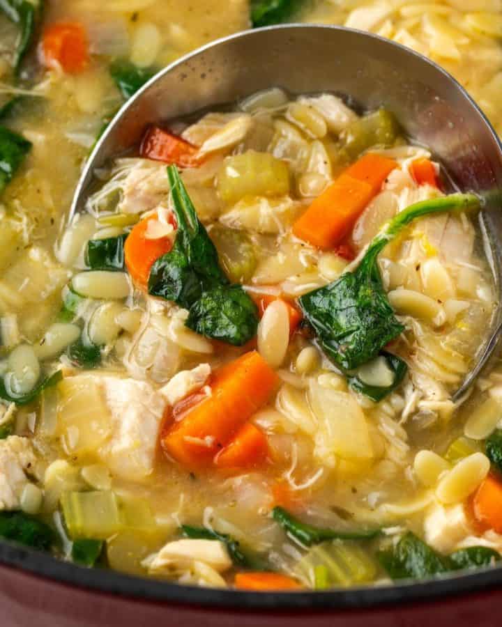 Close up of a silver ladle scooping up lemony chicken orzo soup with spinach from a large pot.