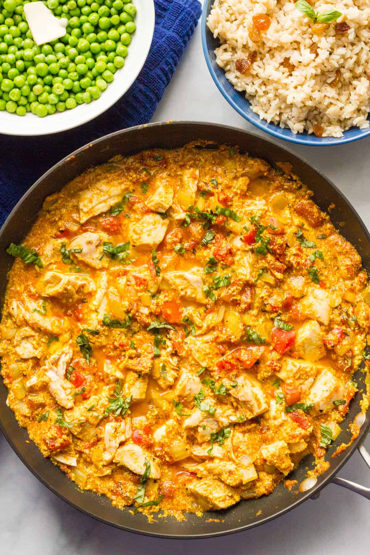 A large skillet of quick chicken curry in a tomato sauce with bowls of rice and green peas nearby.