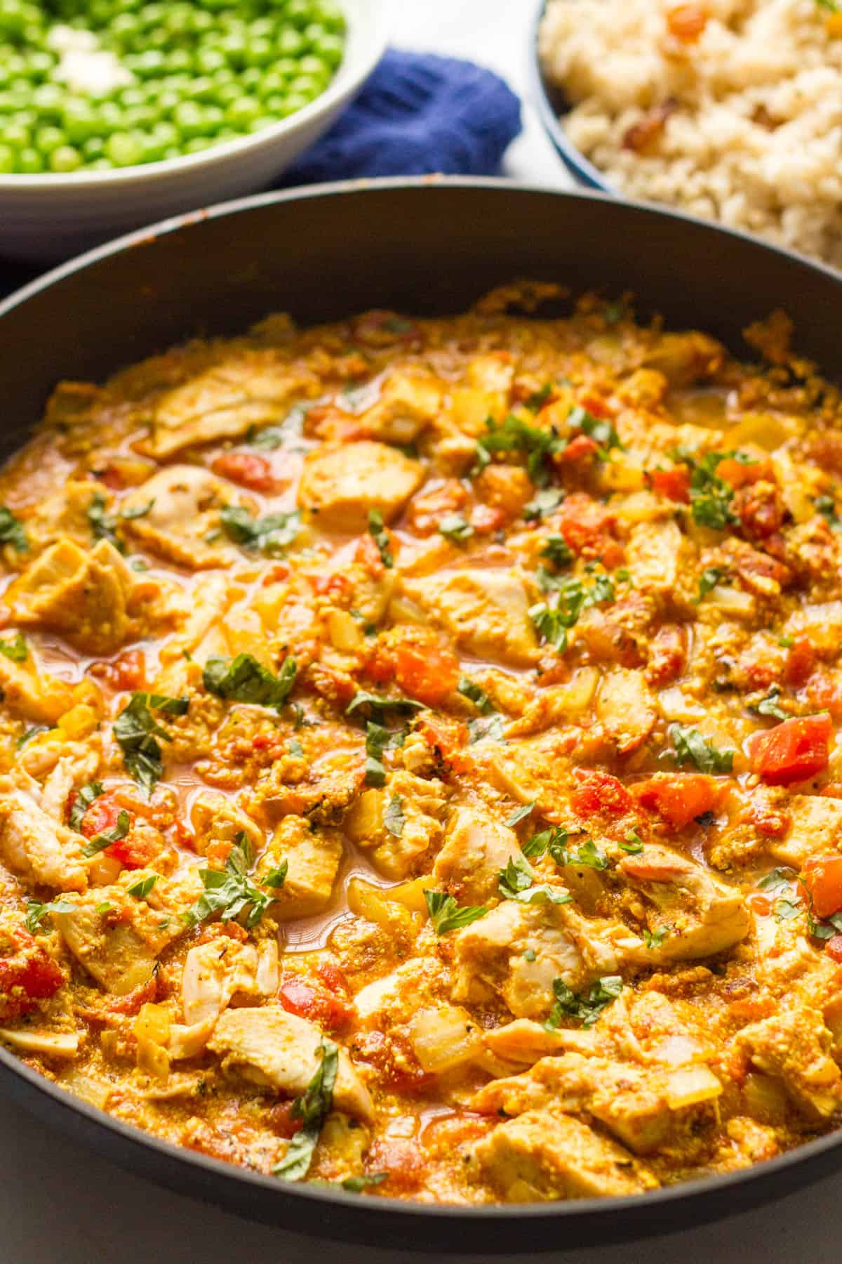 A large dark skillet with cooked chicken curry topped with fresh parsley with bowls of rice and peas in the background.