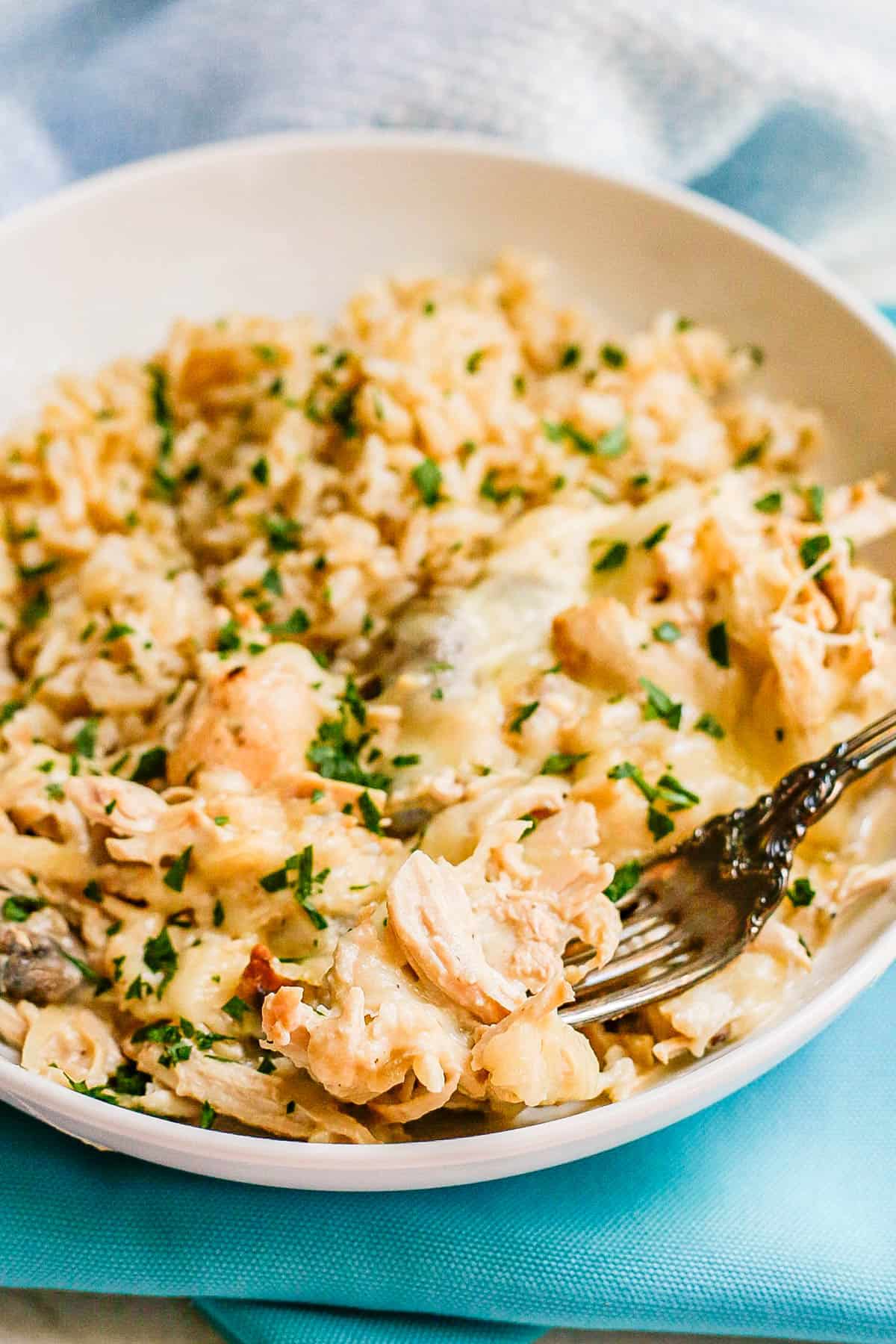 A fork scooping up a bite of creamy cheesy chicken from a white serving bowl with the chicken mixture over rice.