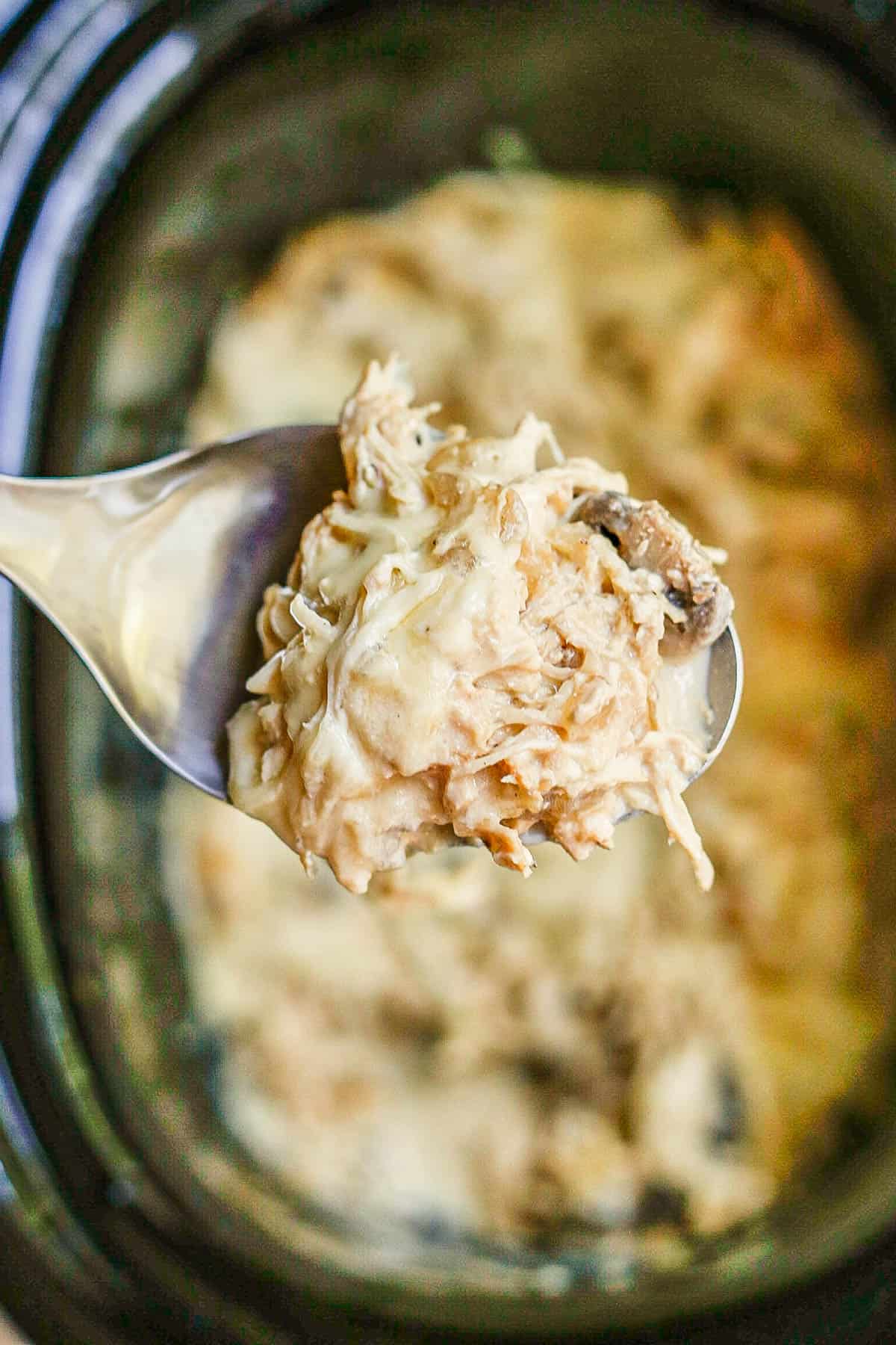 A silver serving spoon scooping up some cheesy cream cheese chicken from a slow cooker insert.