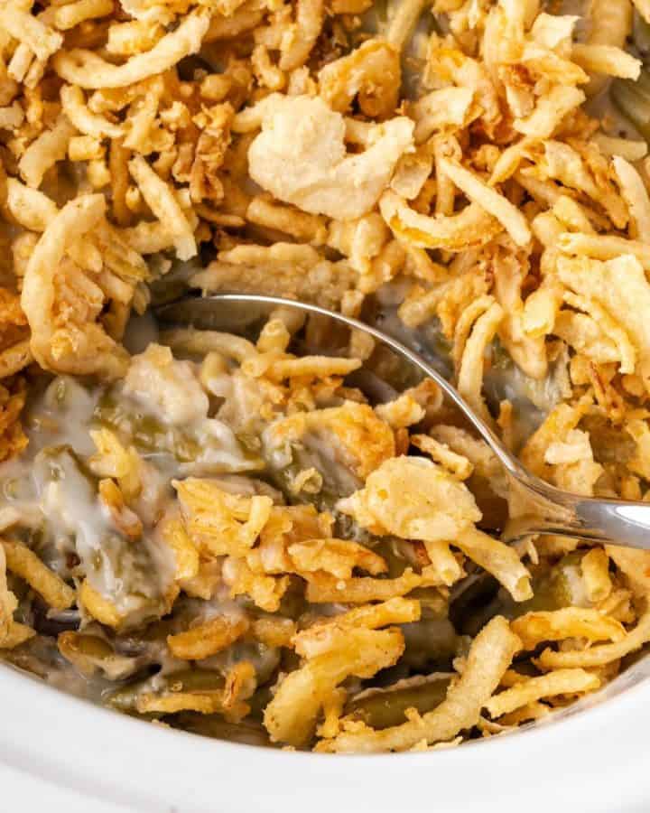 A silver serving spoon scooping up a serving of green bean casserole from a white crock pot.